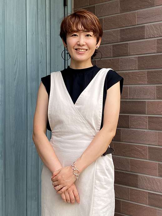 A woman wearing a white dress and black shirt, smiling while standing in front of a blue door and brick wall background.