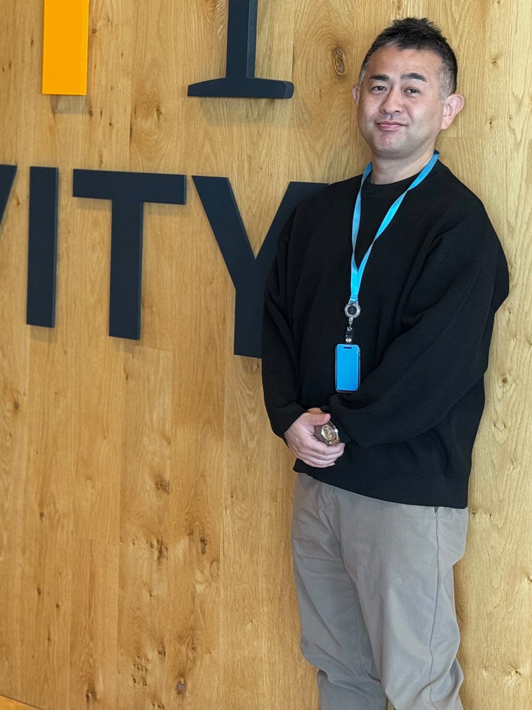 A man stands in front of a wooden wall with large text, wearing a black sweater, khaki pants, and an office badge on a lanyard. The image appears to be taken in an office environment.