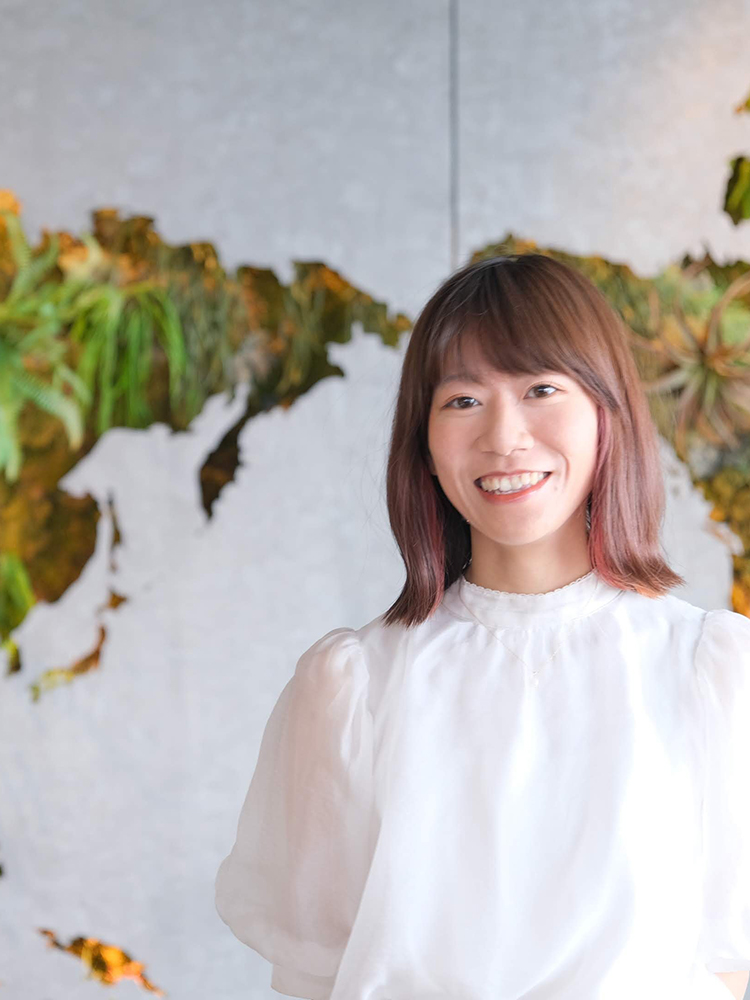 A smiling woman wearing a white blouse stands in front of a green wall background with a map design.