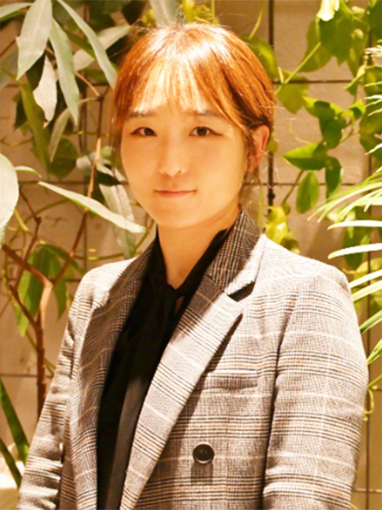 A woman wearing a plaid blazer standing in front of a background filled with indoor plants.