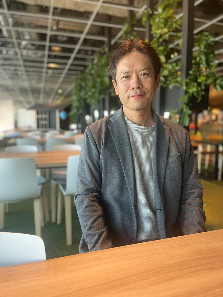 A man sitting at a wooden table in a modern cafe, wearing a casual blazer and light sweater. The background features contemporary decor with green plants and light-colored chairs.