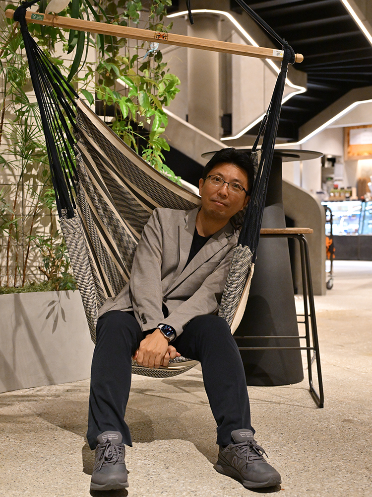 A man in casual attire sits comfortably in a hammock chair within a modern indoor environment featuring greenery and contemporary decor.