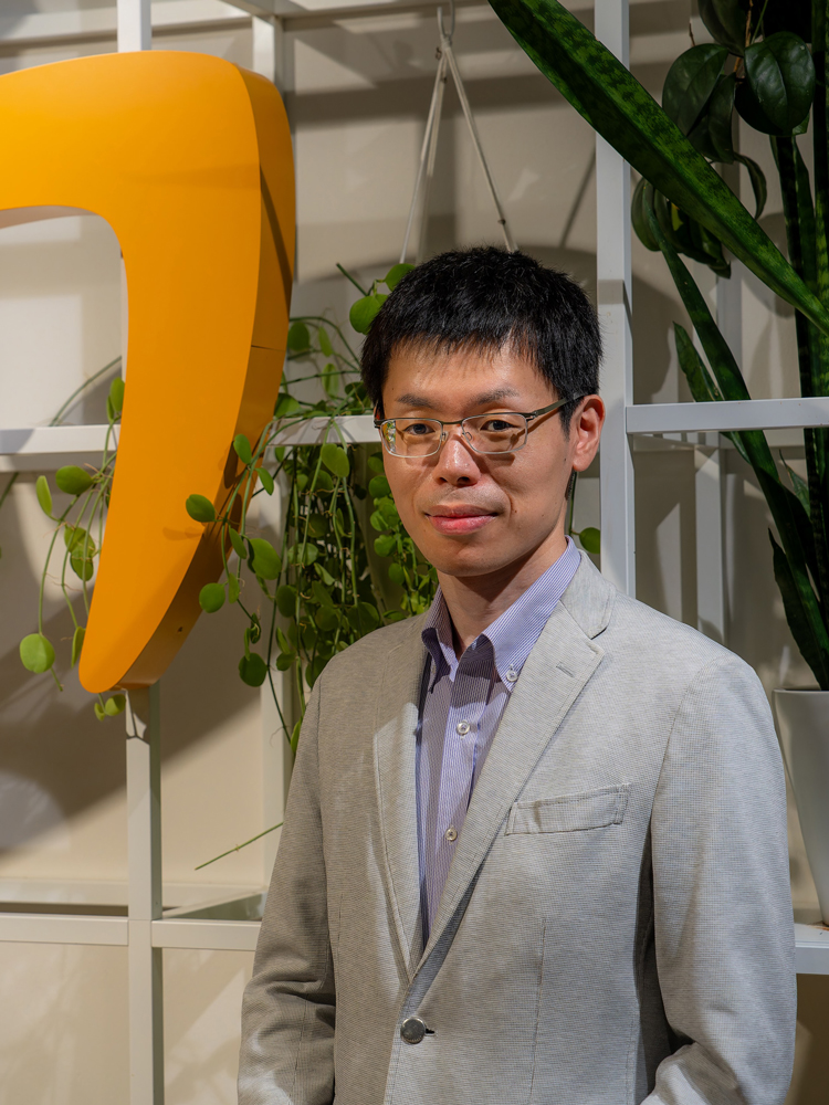 A man in a light gray suit and glasses stands indoors in front of green plants and a large yellow structure.