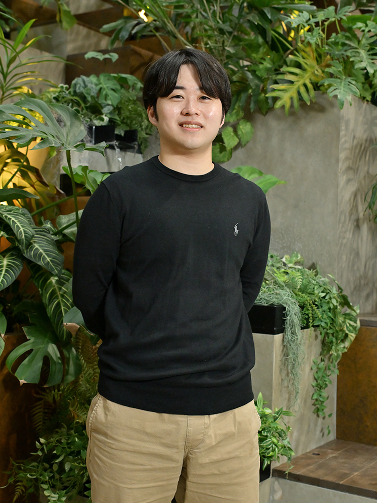 A man wearing a black sweater and beige pants standing in front of a background filled with various indoor plants.