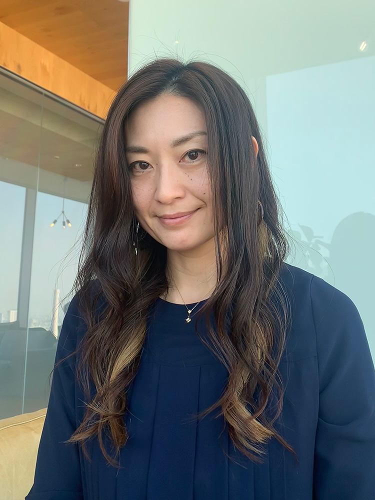 A woman with long wavy hair wearing a navy top, smiling in a modern office setting with large windows and contemporary furnishings.