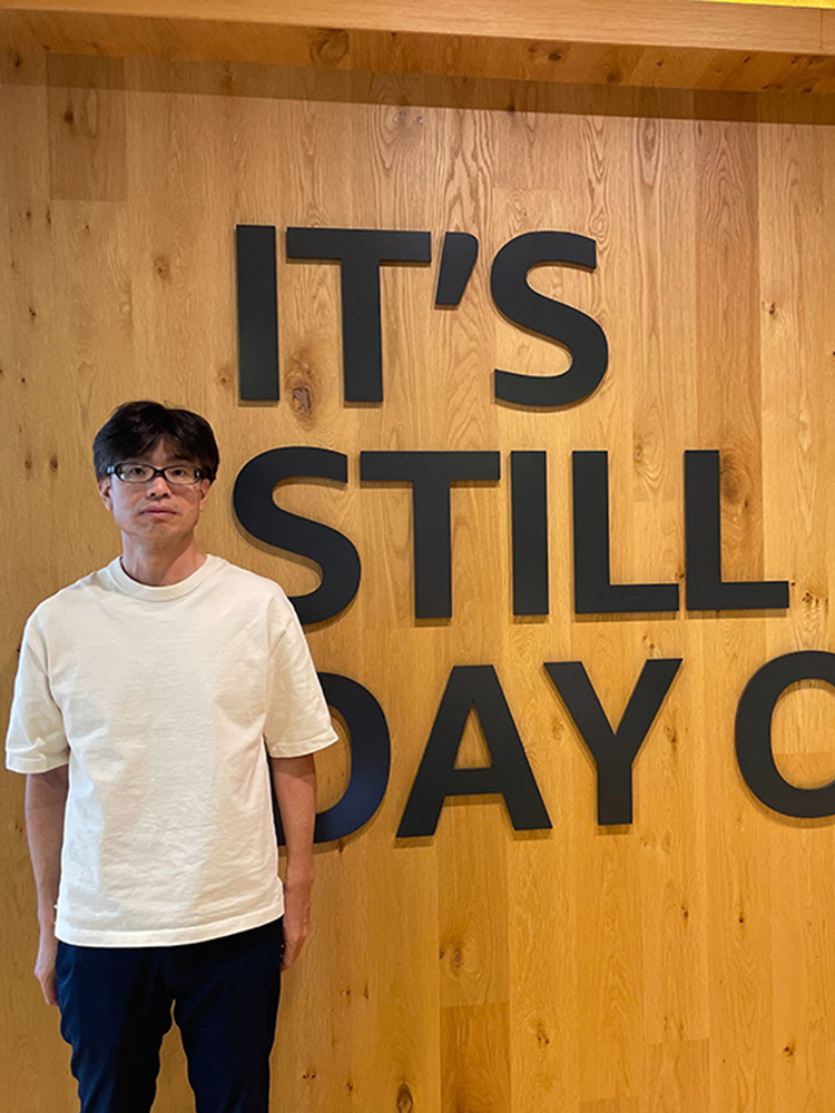A man in a white shirt stands in front of a wooden wall with large text that reads 'IT'S STILL DAY ONE'.