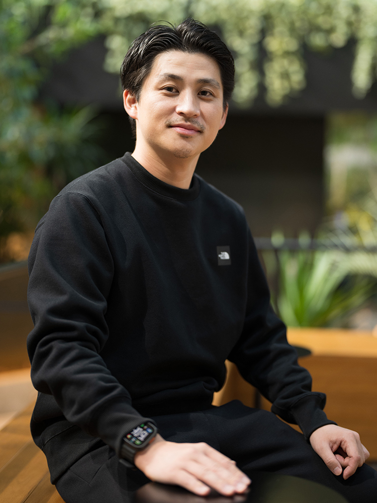 A man wearing a black sweatshirt is seated indoors, looking at the camera with a slight smile. The background features greenery and soft lighting.