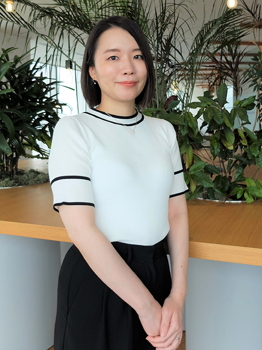 A woman is standing in an office environment with various green plants in the background. She is dressed in business attire and smiling, conveying a professional atmosphere.