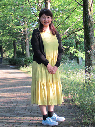 A woman wearing a yellow dress and black cardigan standing on a park pathway, surrounded by green trees and grass, in an outdoor setting.