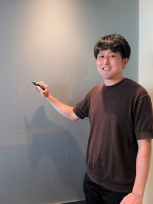 A person in casual attire is smiling and pointing at a whiteboard with a pen in hand.