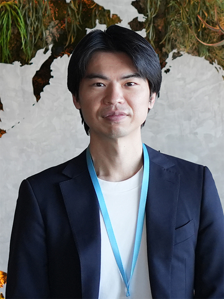 A portrait of a businessman wearing a dark blazer and a white shirt with a blue lanyard, posing indoors against a background featuring a world map design and greenery.
