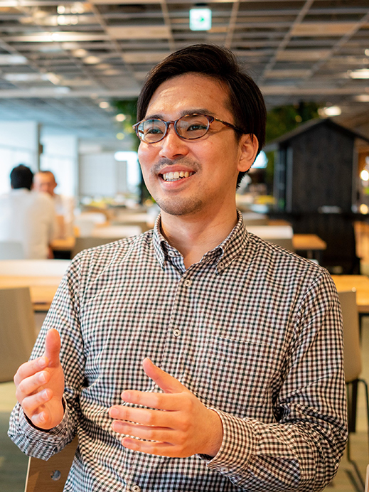 A smiling man wearing glasses and a checkered shirt is speaking in a casual office environment, with tables and colleagues in the background.