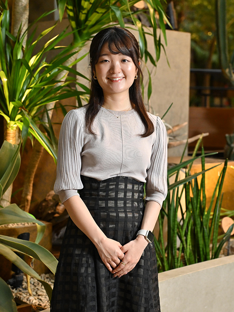 A woman stands smiling indoors in front of a lush garden background, wearing a light top and a checked skirt.