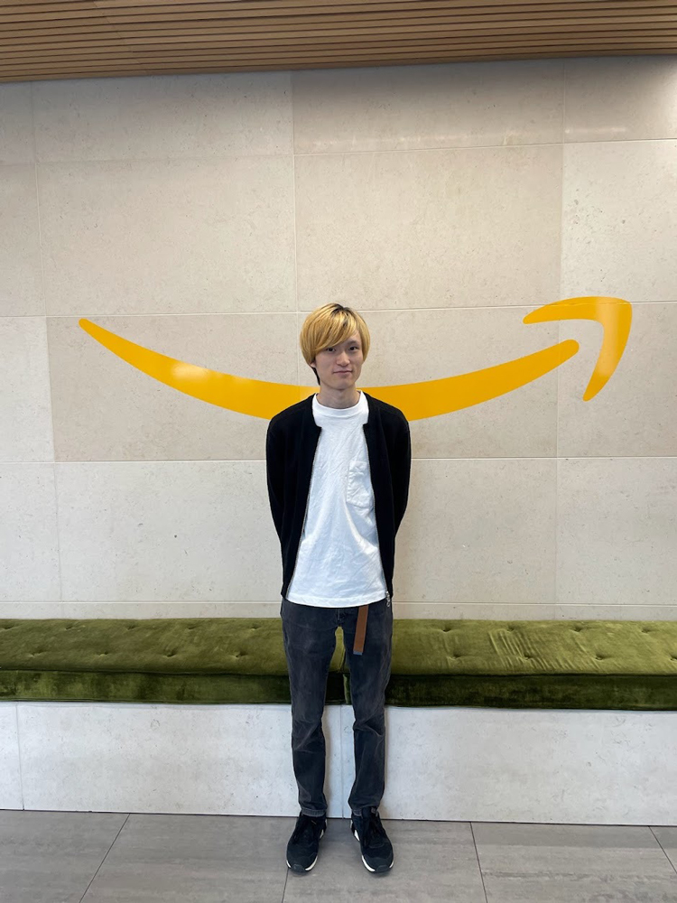 A person standing in front of a wall featuring a large yellow Amazon logo, inside a modern indoor space with a green bench.