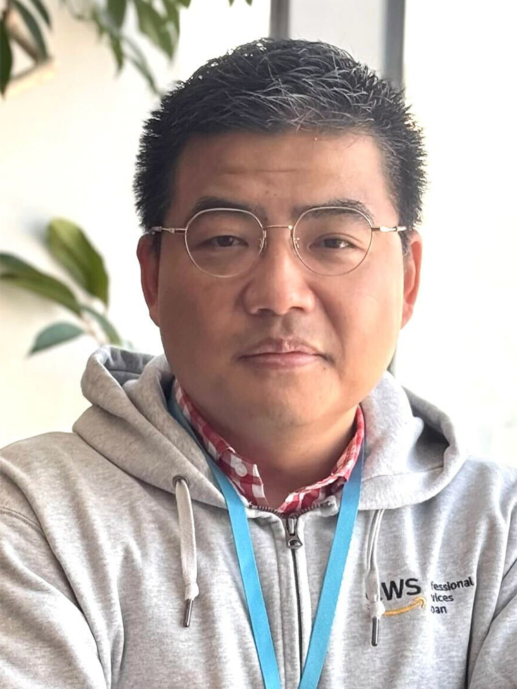 A portrait of an AWS Professional Services employee wearing glasses and a grey AWS hoodie, sitting indoors with plants and natural light in the background.