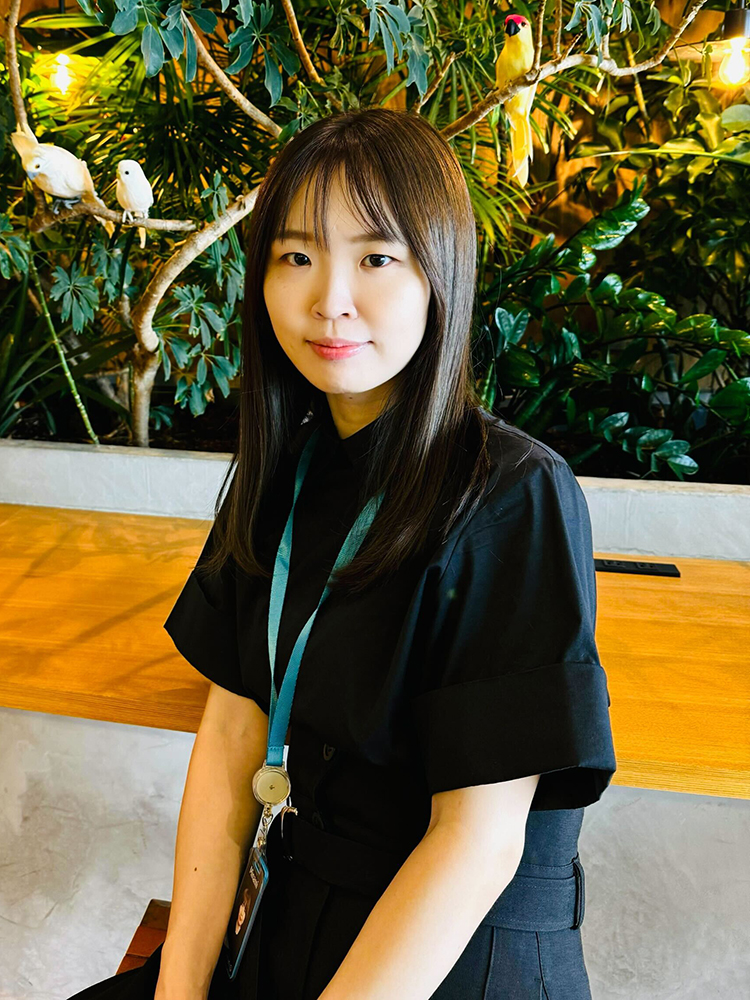 A woman sitting at a café table with artificial birds perched on branches and lush greenery in the background.