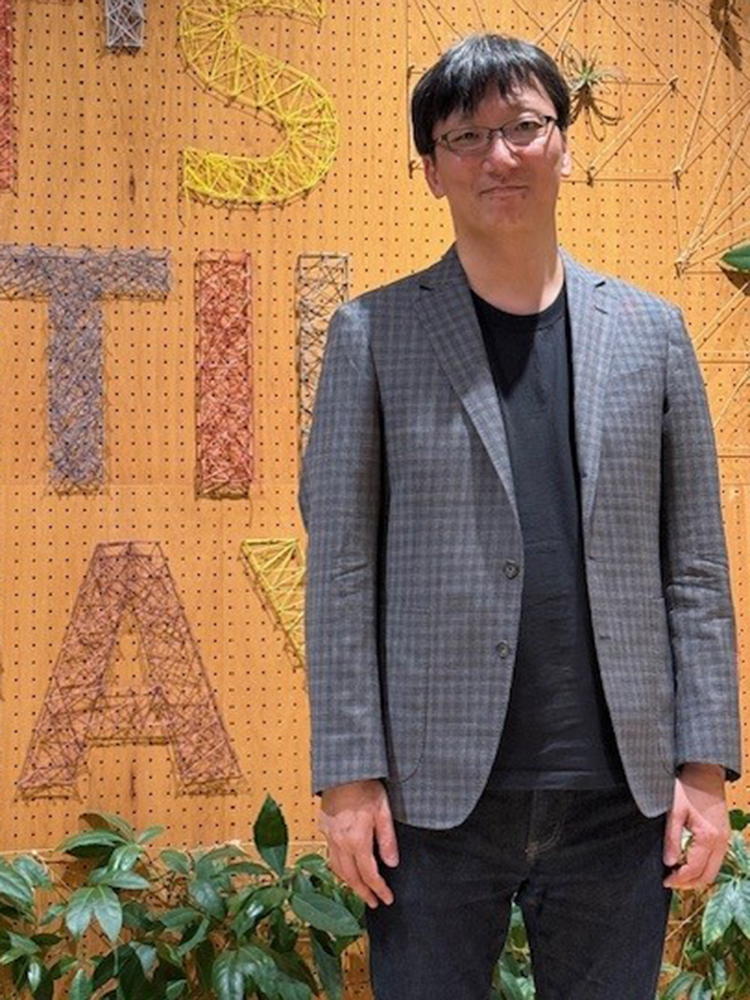 A man wearing a gray checked blazer and black shirt stands in front of a wall decorated with colorful string art letters and geometric shapes. Green plants are visible at the bottom of the image.