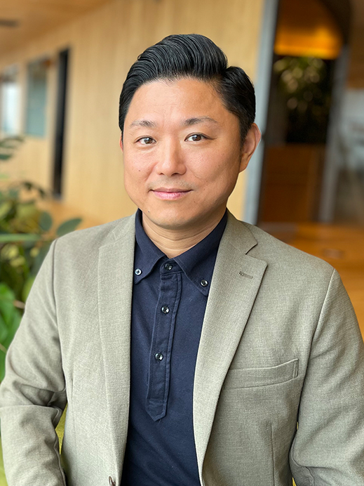 A portrait of a businessman wearing a beige blazer and navy shirt, standing in a modern office environment with plants and wooden walls in the background.