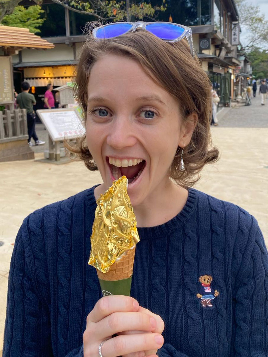 A woman smiling and about to eat an ice cream cone covered with a sheet of edible gold leaf, standing outdoors in a casual setting.