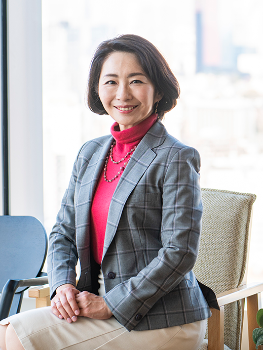 A professional woman sits and smiles in an office chair by a window, dressed in business attire with a cityscape in the background.