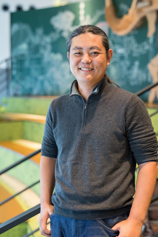 A man with long hair, wearing a dark sweater and jeans, is standing and smiling in a modern office environment with green steps and a chalkboard-style wall in the background.