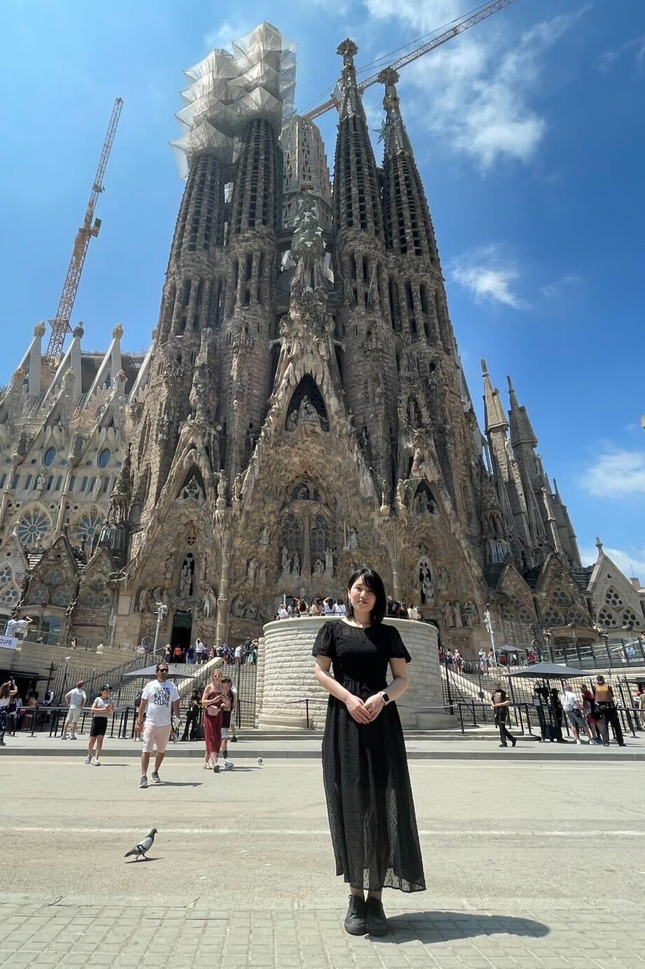 A woman in a black dress stands in front of the Sagrada Familia, a famous basilica in Barcelona, Spain, on a sunny day.