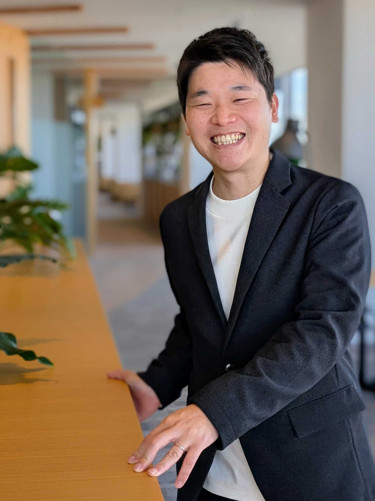 A person in business attire is smiling while standing in a modern office setting, with natural light and plants visible in the background.