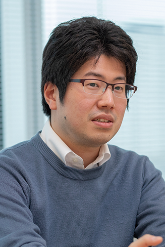 Portrait of a man with glasses in a blue sweater and white shirt, seated indoors, photographed for a DENSO solution case study.