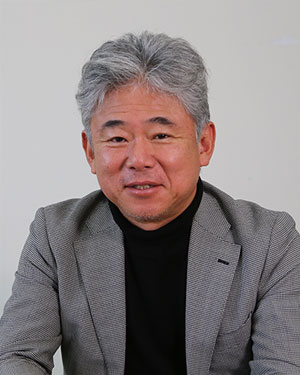A person with gray hair wearing a gray blazer and black shirt against a plain background.