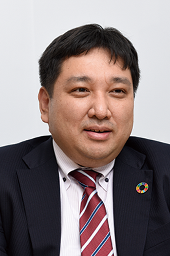 A businessman wearing a dark suit and a striped tie, featuring a Sustainable Development Goals (SDGs) pin on his lapel, shown from the chest up.