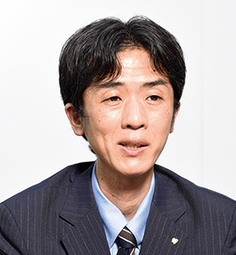A man wearing a dark business suit, blue shirt, and tie, facing forward in a professional portrait setting.