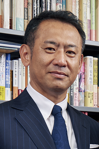 A portrait of a man in a suit and tie standing in front of a bookshelf filled with books.