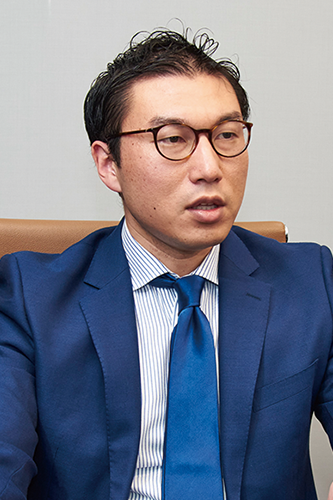A business professional wearing a blue suit, glasses, and a blue tie is seated and speaking.