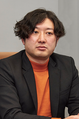 A man with medium-length dark hair, wearing a black jacket and an orange sweater, is seated and looking slightly to the side against a neutral background.
