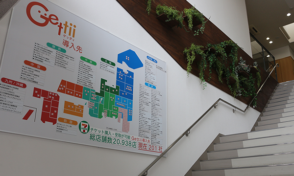 A photo of an office building stairwell with a large, colorful directory sign for 'Gettii' mounted on the wall. The sign features a layout map and list of offices, with some plants decorating the stairwell area.