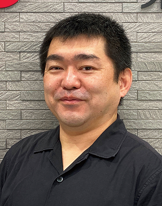 A man with short dark hair wearing a black collared shirt, standing in front of a gray brick wall, facing the camera and smiling slightly.