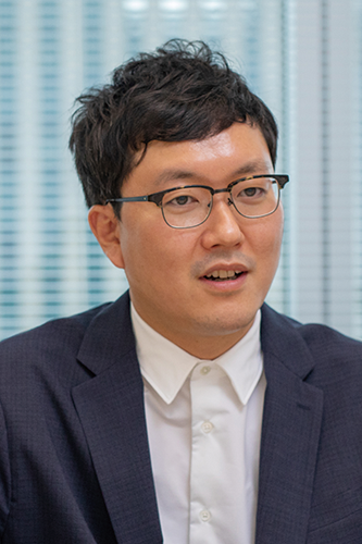 A professional business portrait of a man in a suit and glasses, seated in an office setting, representing DENSO. He has short dark hair and is wearing a white shirt and dark blazer.
