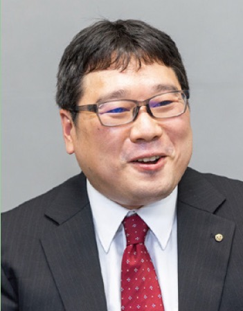 A portrait of a professional man wearing glasses, a business suit, and a red tie, smiling against a plain background.