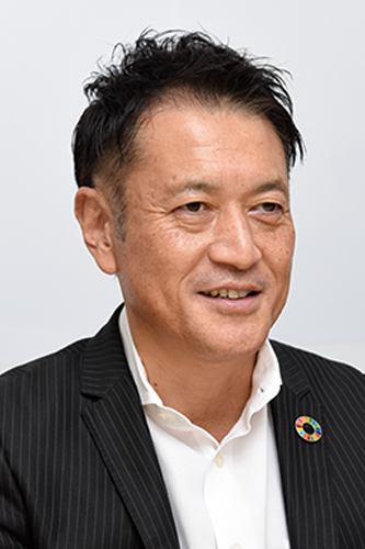 A portrait of a businessman wearing a pinstripe suit and white shirt, featuring an SDGs (Sustainable Development Goals) pin on his lapel.