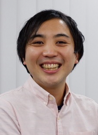 A person smiling and wearing a light pink shirt, photographed indoors in front of a light background.