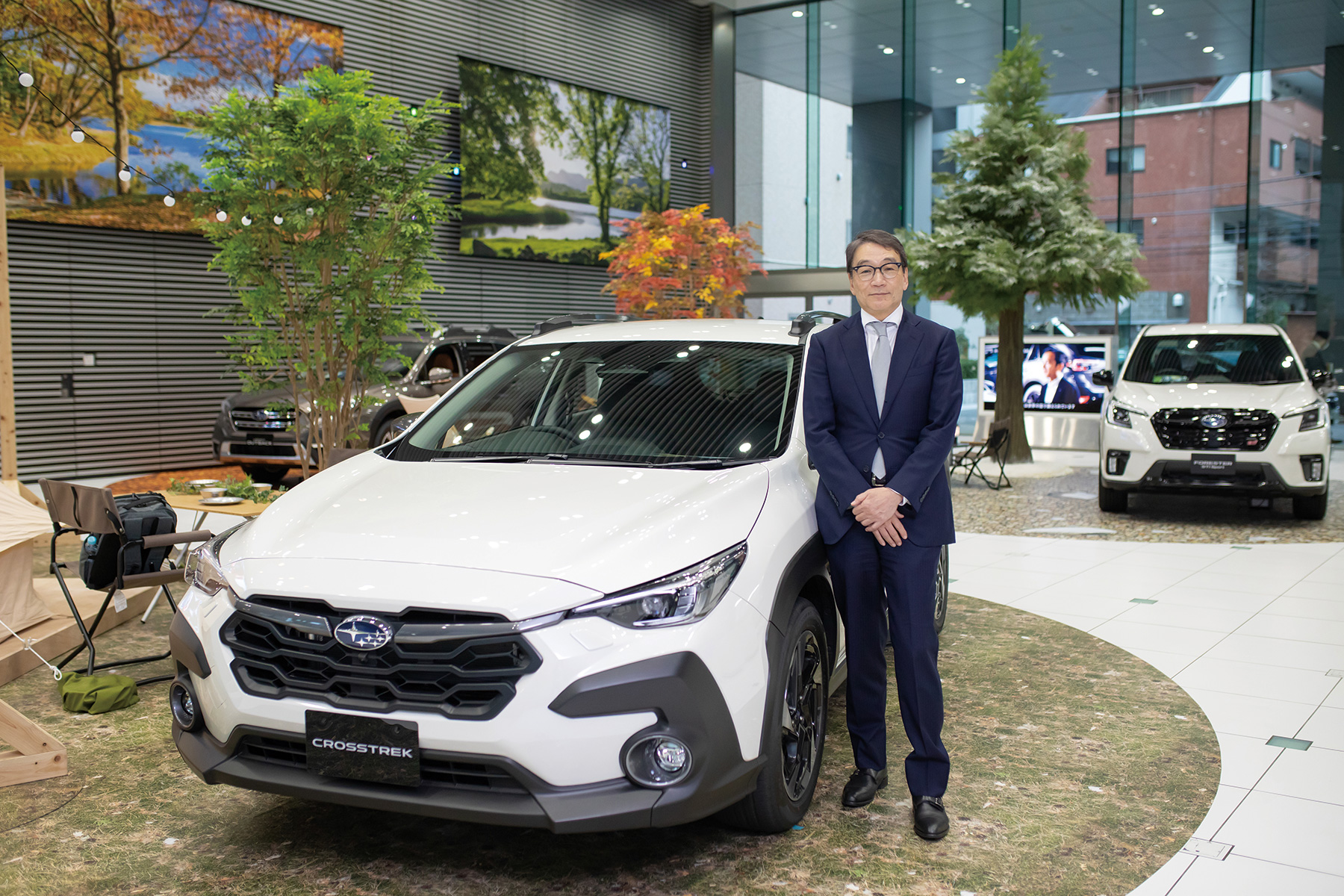 A white Subaru Crosstrek displayed in a showroom with nature-themed decor and other vehicles in the background.