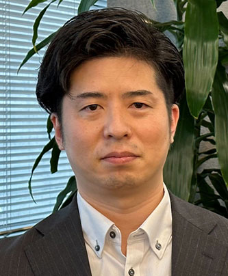 Portrait photograph of Masahide Tomie in a business suit, featured in the Toda Kensetsu case study. The person is seen against an office background with blinds and plants.