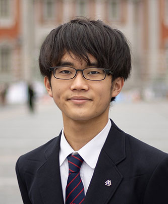 A young man wearing glasses, a dark suit, white shirt, and striped tie standing outdoors with a blurred building in the background.