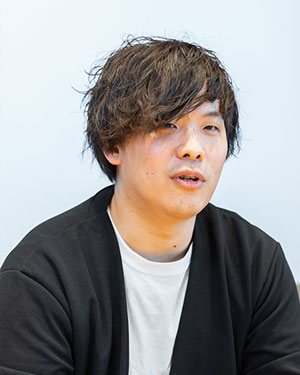 A person wearing a black cardigan over a white shirt, with short, slightly messy dark hair, against a light blue background.