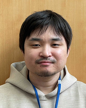 A portrait photograph of a man wearing a hoodie, standing in front of a wooden background.