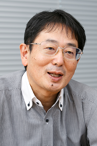 Portrait of an Asahi Kasei employee, featuring a middle-aged man with glasses, wearing a collared shirt, and sitting in front of a striped background.