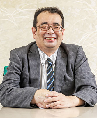 A professional portrait of a man in a business suit and striped tie sitting at a table, smiling, with a neutral background. Used for Nabtesco case study speaker introduction.