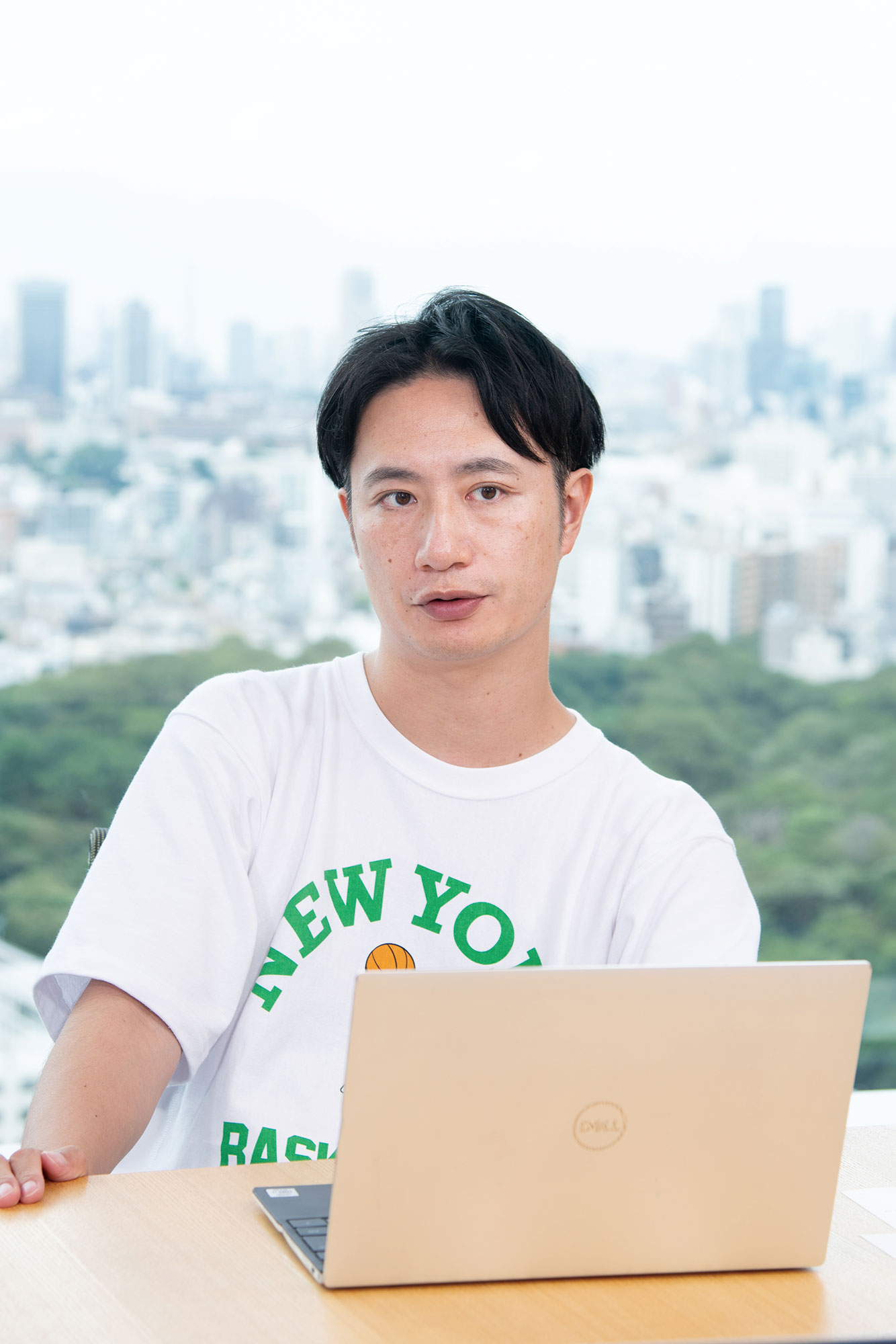 A person wearing a white "New York Basketball" t-shirt sits at a table with a Dell laptop, with a cityscape and greenery visible in the background.