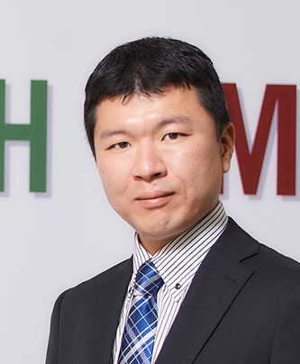 Portrait of Hikaru Konoe, a speaker for Nishiyamato, featured in an AWS case study. The individual is dressed in a business suit, standing in front of a background with partial visible green and red letters.