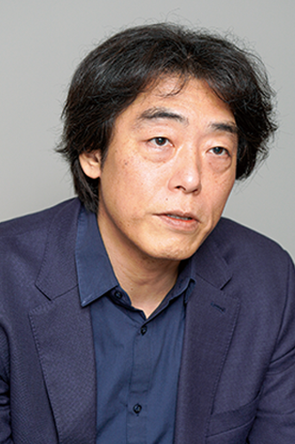 Portrait of an individual with medium-length dark hair wearing a navy suit and shirt, set against a plain gray background.